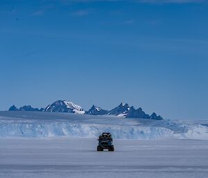 A Hagglund vehicle on sea ice, with dramatic mountain peaks and and ice cliff in the distance.