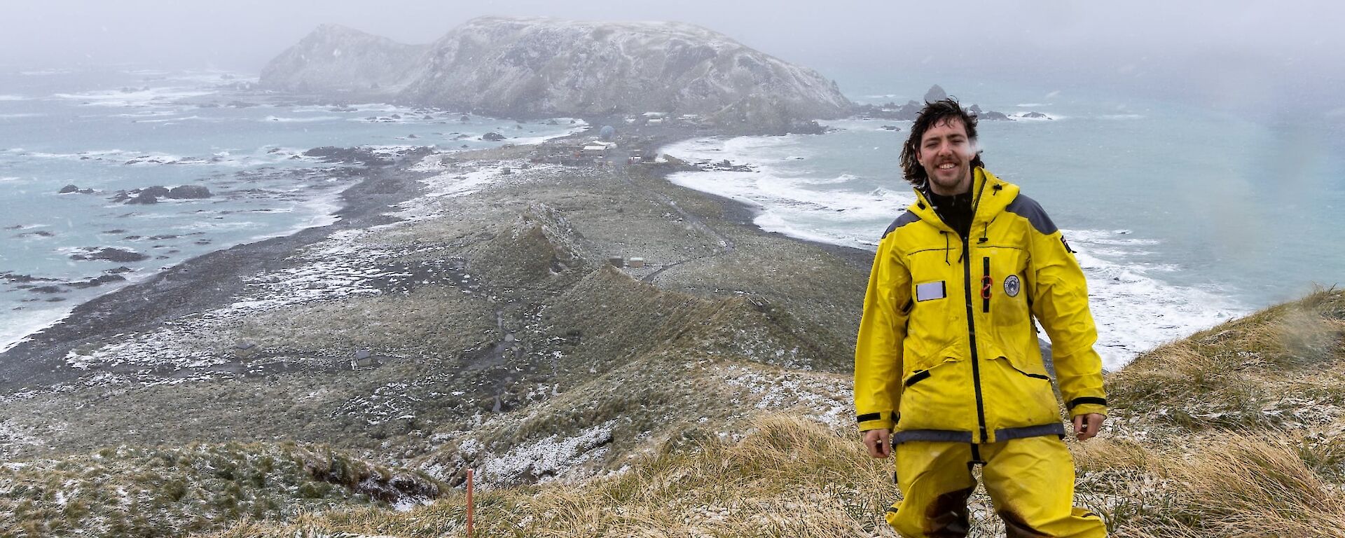 A smiling man wearing yellow coloured field clothing stands atop a grassy hill with a snow and mist covered isthmus and buildings visible in the background.