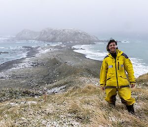 A smiling man wearing yellow coloured field clothing stands atop a grassy hill with a snow and mist covered isthmus and buildings visible in the background.
