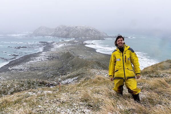 A smiling man wearing yellow coloured field clothing stands atop a grassy hill with a snow and mist covered isthmus and buildings visible in the background.