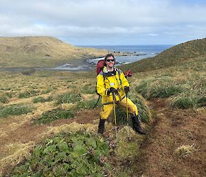 A smiling man in yellow outdoor clothing stands amongst green plants set in a rugged rolling landscape that drops into a bay in the sea in the distance.