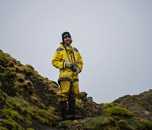 A smiling man in yellow outdoor clothing stands amongst tussock grass and other green plants.