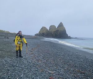 A smiling man in yellow outdoor gear stands on a black pebbly beach. In the distance three striking rock stacks jut from the ocean.