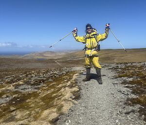 A smiling man in yellow outdoor clothing holds two poles in the air. he is standing on a rocky track in a rolling, tussock covered landscape.