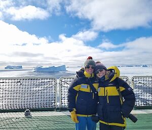 Two female expeditioners on the deck of a ship with icebergs behind them