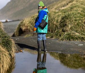 A woman in wellington boots and outdoor gear stands at the edge of a large puddle that reflects her image like a mirror.