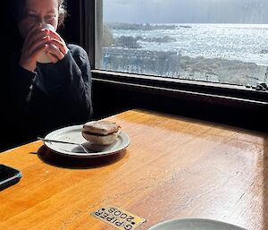 A woman sits at a table by a window, bathed in sunlight, drinking coffee.