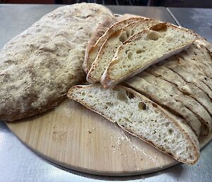 Slices of ciabatta bread next to a whole loaf on a circular wooden chopping board.