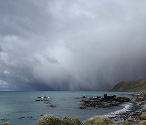 A dark grey sky with looming rain hovers above the flat blue ocean and rugged hills in the distance.
