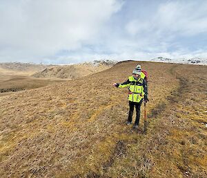 A woman wears a yellow rain jacket and a beanie with a pom pom and stands on a track in a tussock covered landscape with snow-capped hills in the distance.