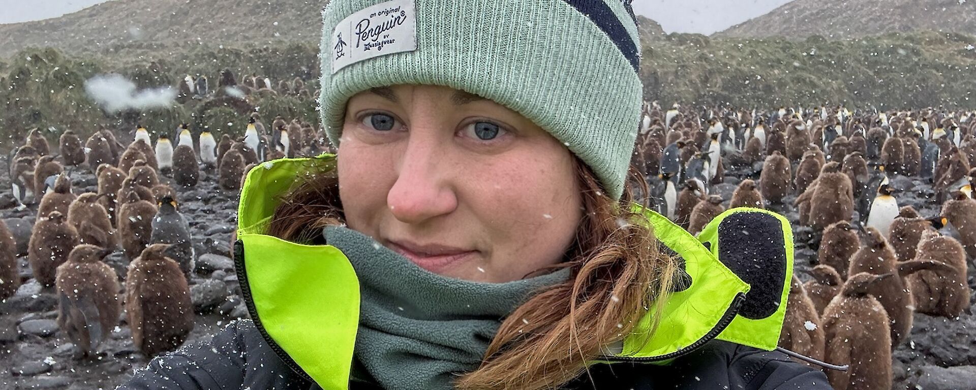 A woman in a beanie stands on a beach in front of brown fluffy King Penguin chicks