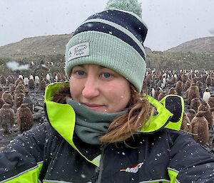 A woman in a beanie stands on a beach in front of brown fluffy King Penguin chicks