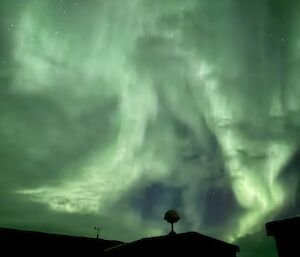 A dazzling green aurora shimmers across the night sky above buildings.
