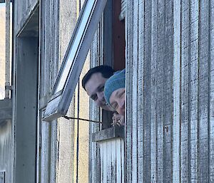 A smiling man and woman peer out of a semi-opened window of a wood clad building.