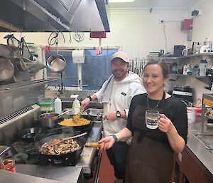 A smiling man and woman stand next to a stove top in a kitchen - both are cooking food in frying pans.