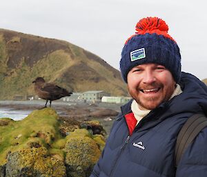 A smiling man wearing a blue and red beanie with a big red pompom stands next to a Skua (bird) that sits on a green, moss-covered rock.