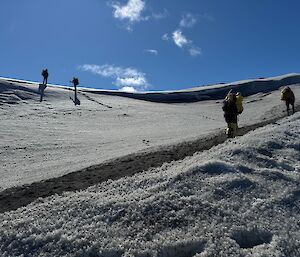 Expeditioners walk up a snow covered slope under a blue sky with a few sparse clouds