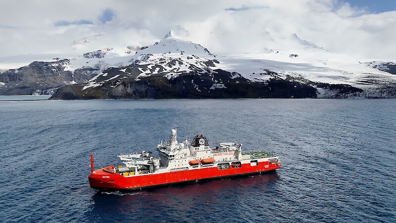 A red and white ship sits in the sea with a snow covered mountainous island behind it