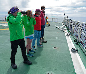 People with binoculars stand on a ship's deck and look out to sea