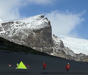 People in bright orange, on a black volcanic plain with rocks and snow behind them and a bright yellow tent
