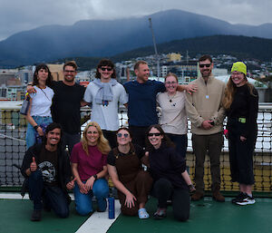 A group of smiling people stand on a ship's deck with kunanyi Mount Wellington behind them