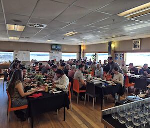 A well lit, open room with three long tables with people sitting along both sides and enjoying a meal