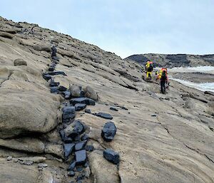 Two expeditioners walk across a rocky landscape with a line of black rocks clearly standing out amongst the brown rocks.