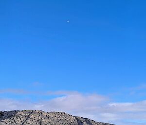 A plane barely visible in the distance flies through a clear blue sky above a rocky outcrop, with sea ice in the foreground.
