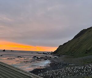 Orange and cloud covered sky as the sun sets over the ocean. There are penguins in the foreground on the beach.