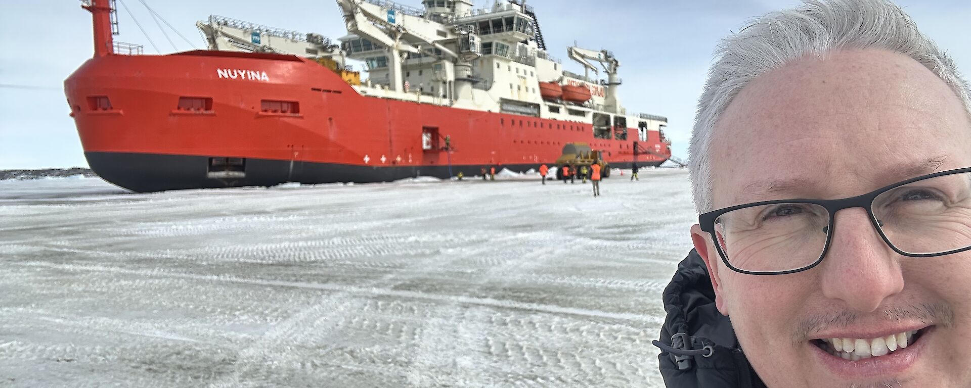 A man looks into the camera, standing on ice, with a red ship in the background