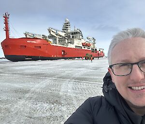 A man looks into the camera, standing on ice, with a red ship in the background