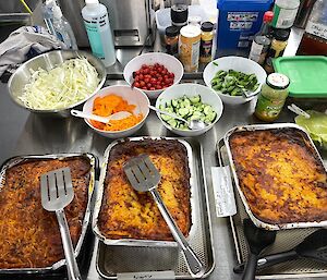 Three foil trays of food with vegetables including tomatoes, cucumbers and cabbage.