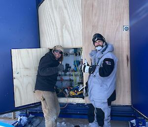 Two men smiling near a switchboard in a trailer