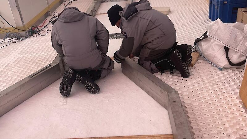 Two peope crouching over a frame that is wide at the bottom and narrow at the top