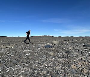 An expeditioner dressed in all black with a white baseball cap runs from left to right along a road in a rocky landscape