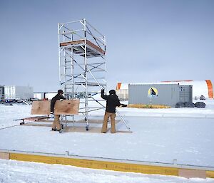Two people on a snowy slope with a metal structure.