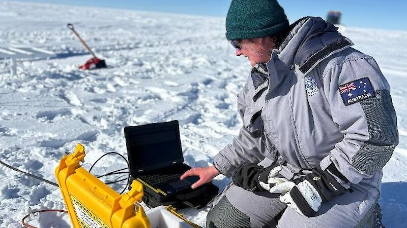 woman checking a laptop connected to an instrument. The woman is kneeling on the snow surface beside the instrument in a yellow box, while typing on the laptop keyboard to her right.