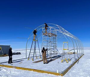 The steel framework for a cylindrical shelter being erected by four people on scaffolding and ladders.