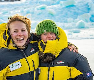 Two women, one with her arm around the other, in yellow puffy jackets, with sea ice behind.