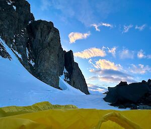 View of steep, rocky cliffs, snow and sky, from inside a yellow bivvy bag.
