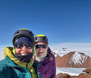 A selfie of two women on top of a mountain, with views to other mountain peaks and ice behind.