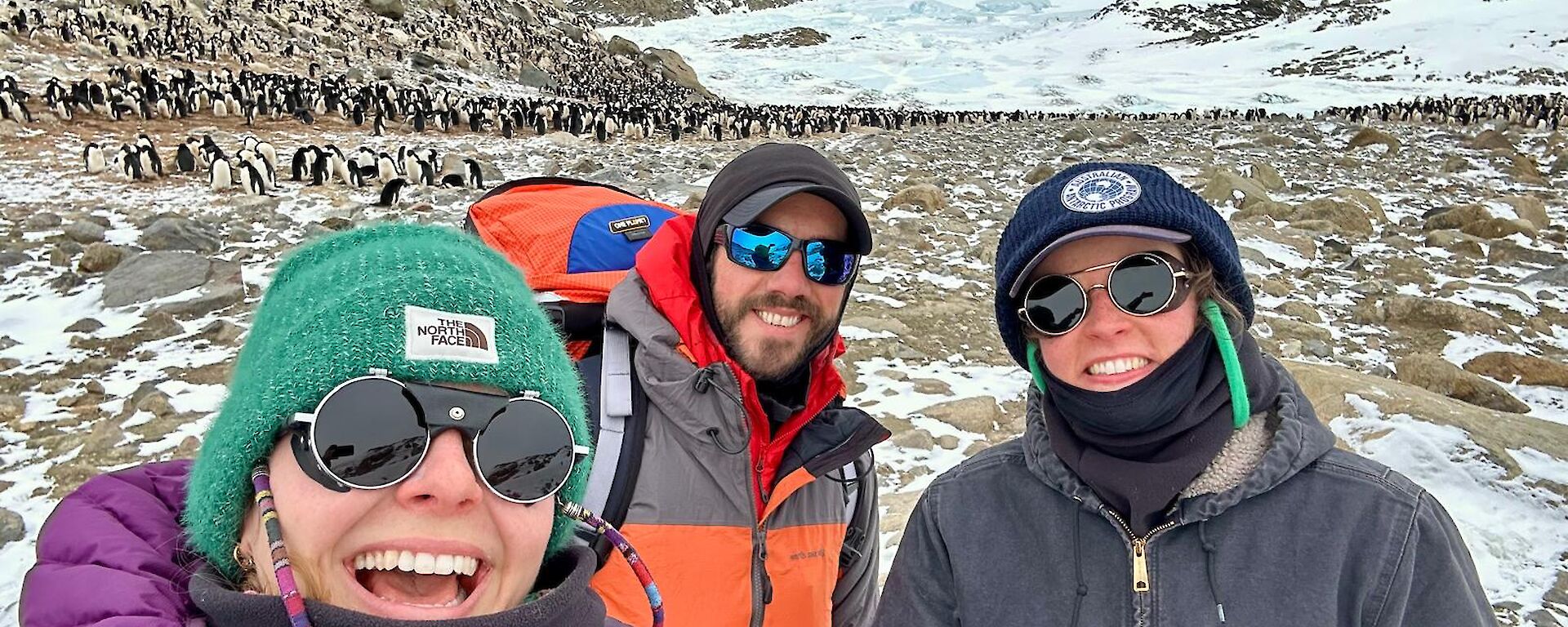 Three people standing in front of an Adelie penguin colony.
