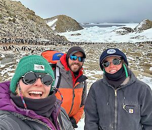 Three people standing in front of an Adelie penguin colony.