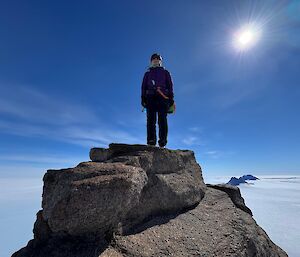 A woman standing on the rocky pinnacle at the top of a mountain, with views of ice behind.