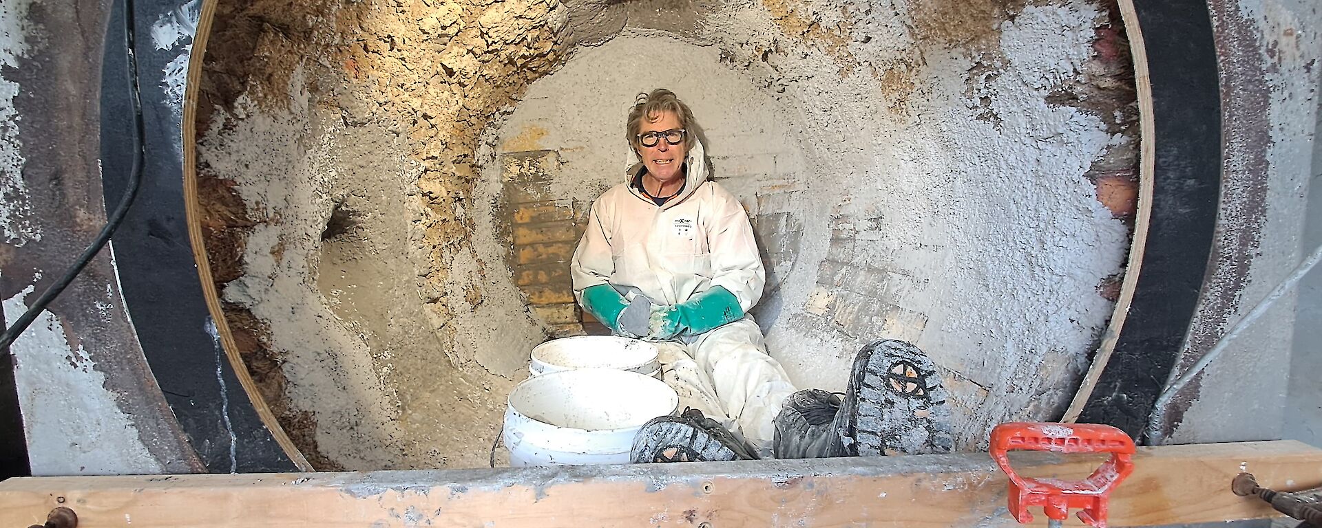 A smiling man wearing a white PPE suit, glasses and gloves sits inside a large round incinerator surrounded by buckets of render.