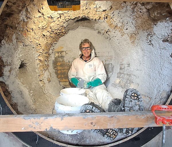 A smiling man wearing a white PPE suit, glasses and gloves sits inside a large round incinerator surrounded by buckets of render.