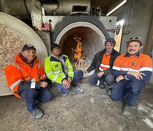 Four smiling men wearing high vis PPE crouch around the open door of a newly renovated incinerator - there is a wooden albatross burning inside.