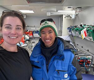 Two people standing in a shipping container surrounded by bags and containers of medical supplies, neatly arranged on shelves.
