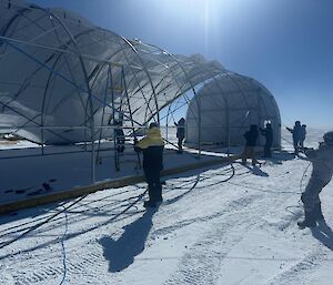 Eight people pull a tarpualin over the steel frame of a cylinder-shaped structure.