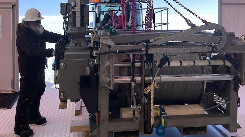 A man in a tractor uses a crane to lower a heavy winch into position inside the million year ice core drill shelter, while another man ensures the winch is in the correct position.